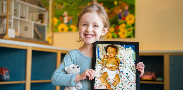 Image of 6-year-old Olivia, now healthy and smiling, holding a photograph of herself as a hospitalized and intubated infant